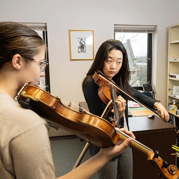 An Asian female professor with black shirt plays a violin while looking at sheet music as a White female student follows along.